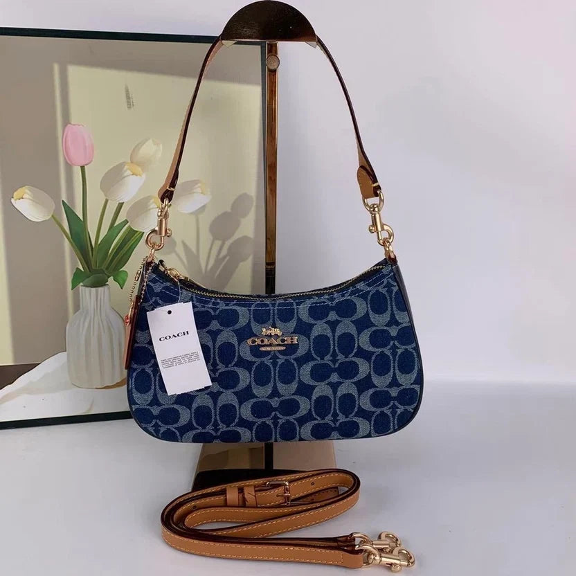Blue Coach handbag with a brown strap on a white surface, next to a vase with flowers.
